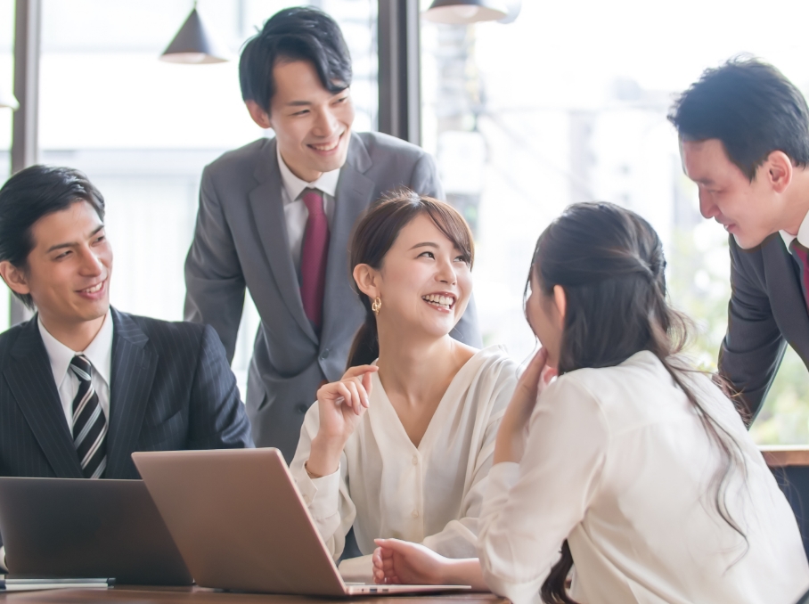 経営課題解決のイメージ。会議室で話し合うビジネススーツ姿の若い男女5人のグループ。笑顔で会話を交わしており、ラップトップが机上に置かれている。