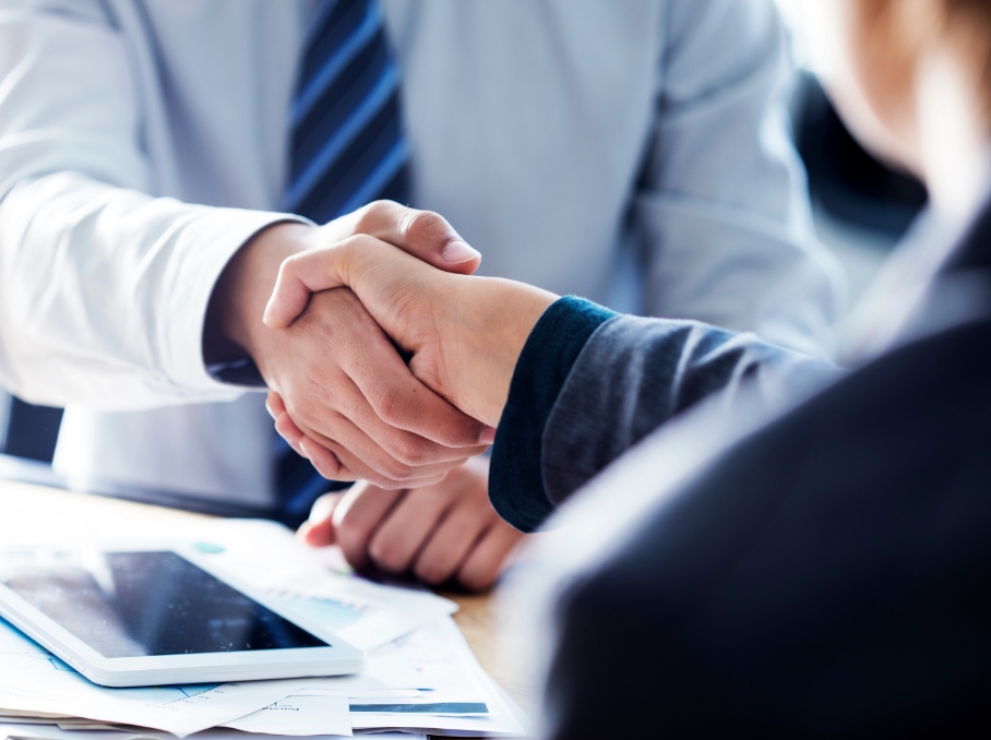 信頼できる出会いのイメージ。ビジネススーツを着た2人の人物が握手しているクローズアップ。デスクには書類とタブレットが置かれている。