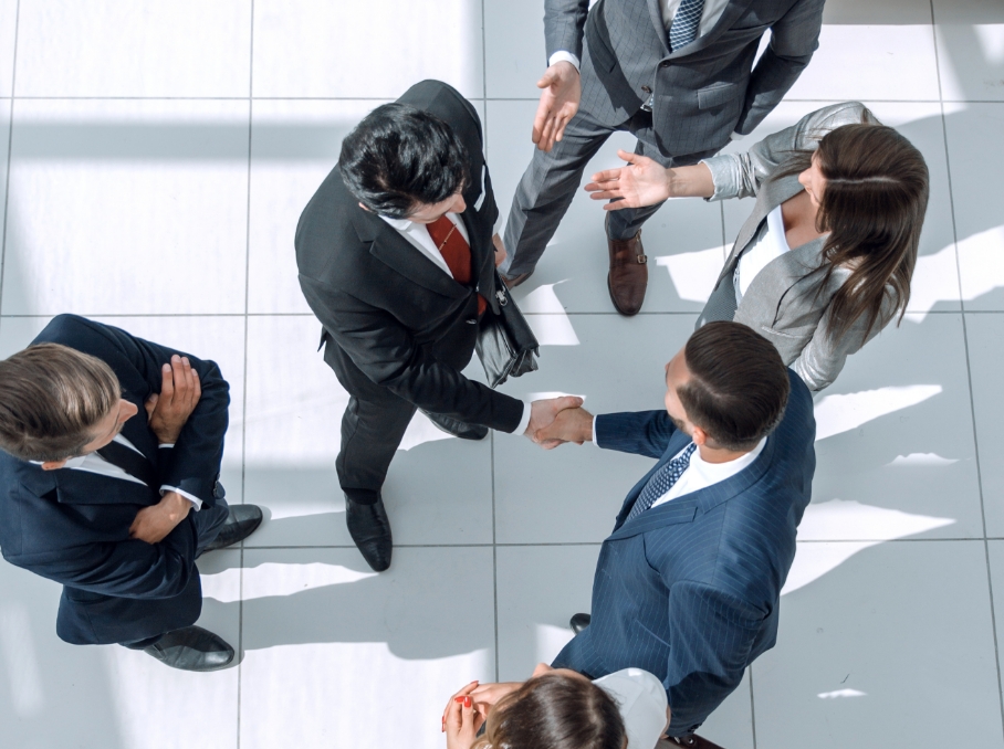 本業に集中のイメージ。屋内の明るいスペースで握手を交わすビジネスパーソンたちを上から見た構図。周囲には会話をしている他の人物もいる。